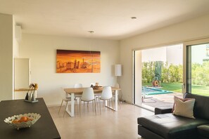 Ein modernes Wohnzimmer mit Essbereich, einer schwarzen Ledersofa, einem Esstisch mit weißen Stühlen, einem Bild an der Wand und Blick ins Freie.