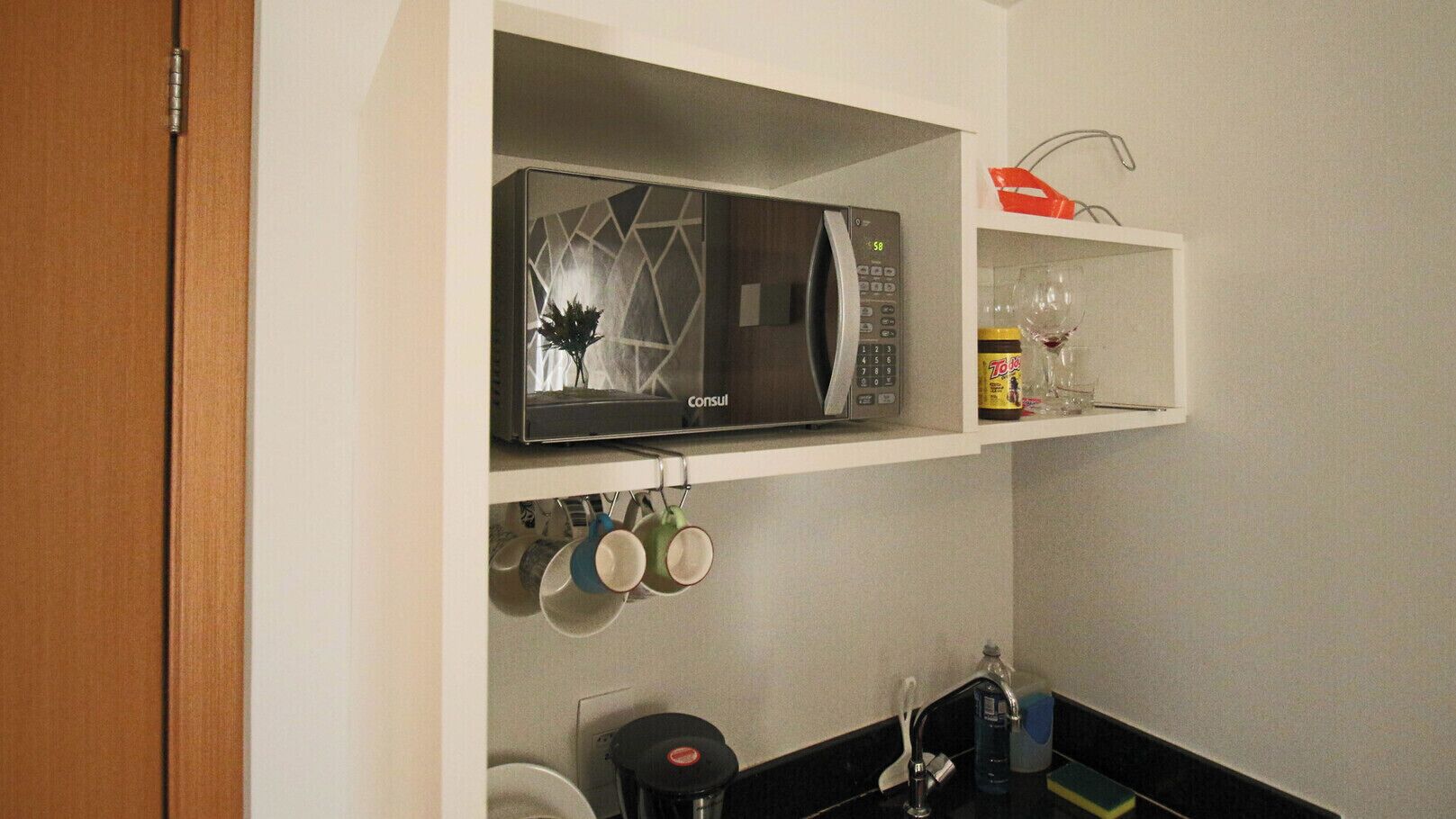 Una cocina con microondas, repisa con un tarro de miel y tazas colgando.