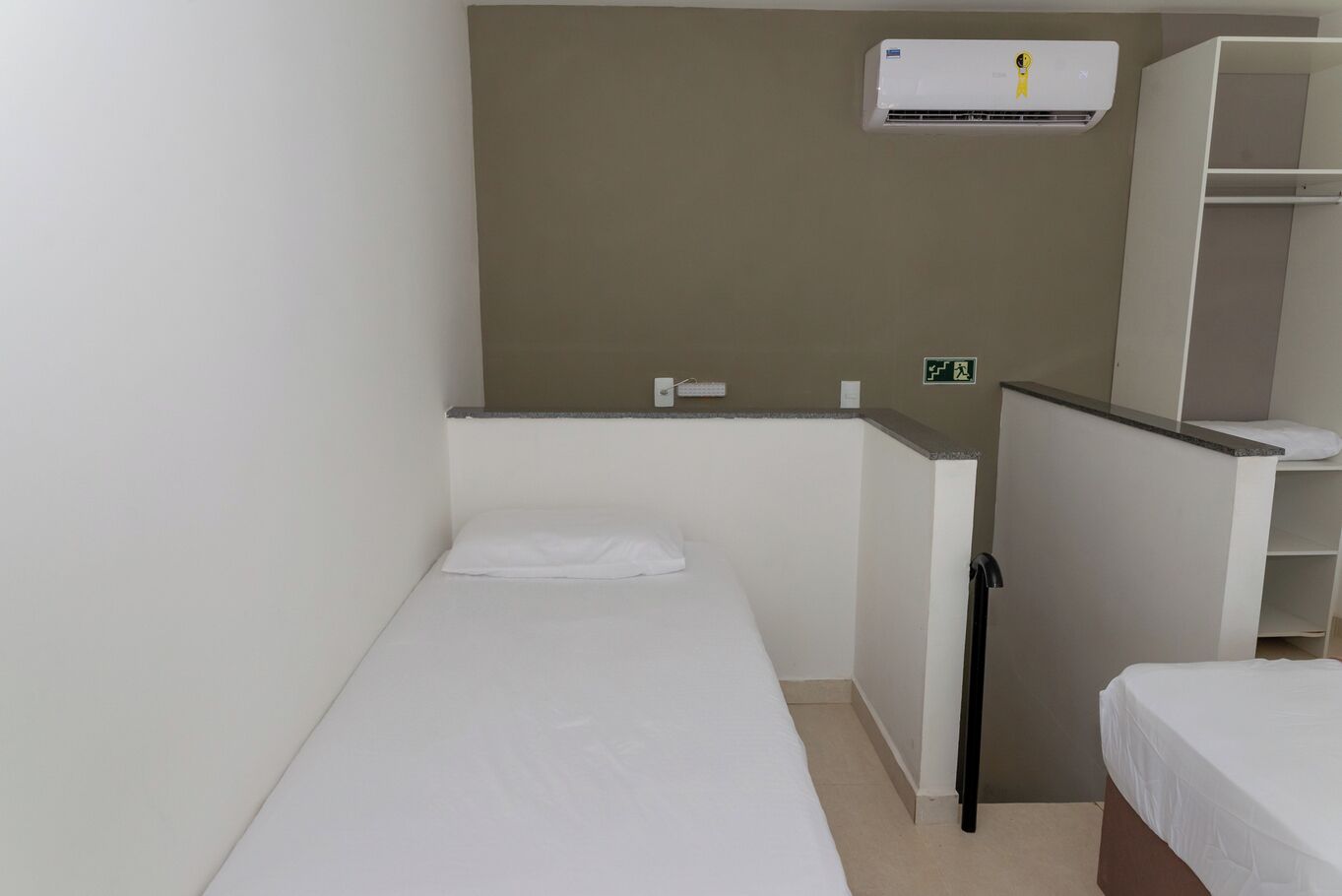 A small, single-bed room with a white bed, a small desk, a chair, a wardrobe, and a wall-mounted air conditioner.