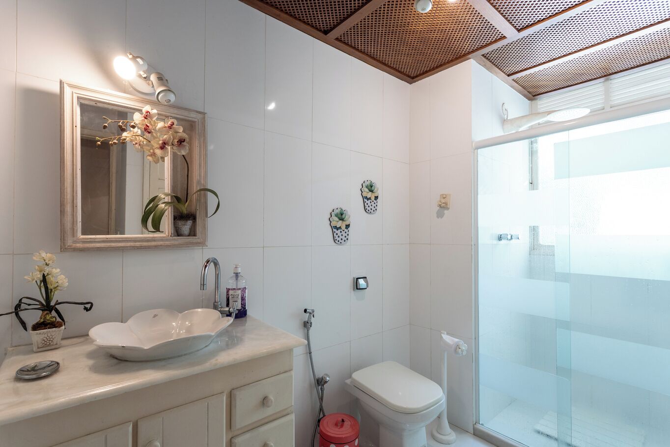 Un bany modern amb un gran mirall, rajoles blanques, un cubicle de dutxa amb vidre i un lavabo blanc amb un ram de flors.