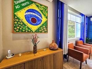 Ein Empfangsbereich mit Holztresen, einem Gemälde der brasilianischen Flagge, einer Vase mit Blumen und orangefarbenen Stühlen.