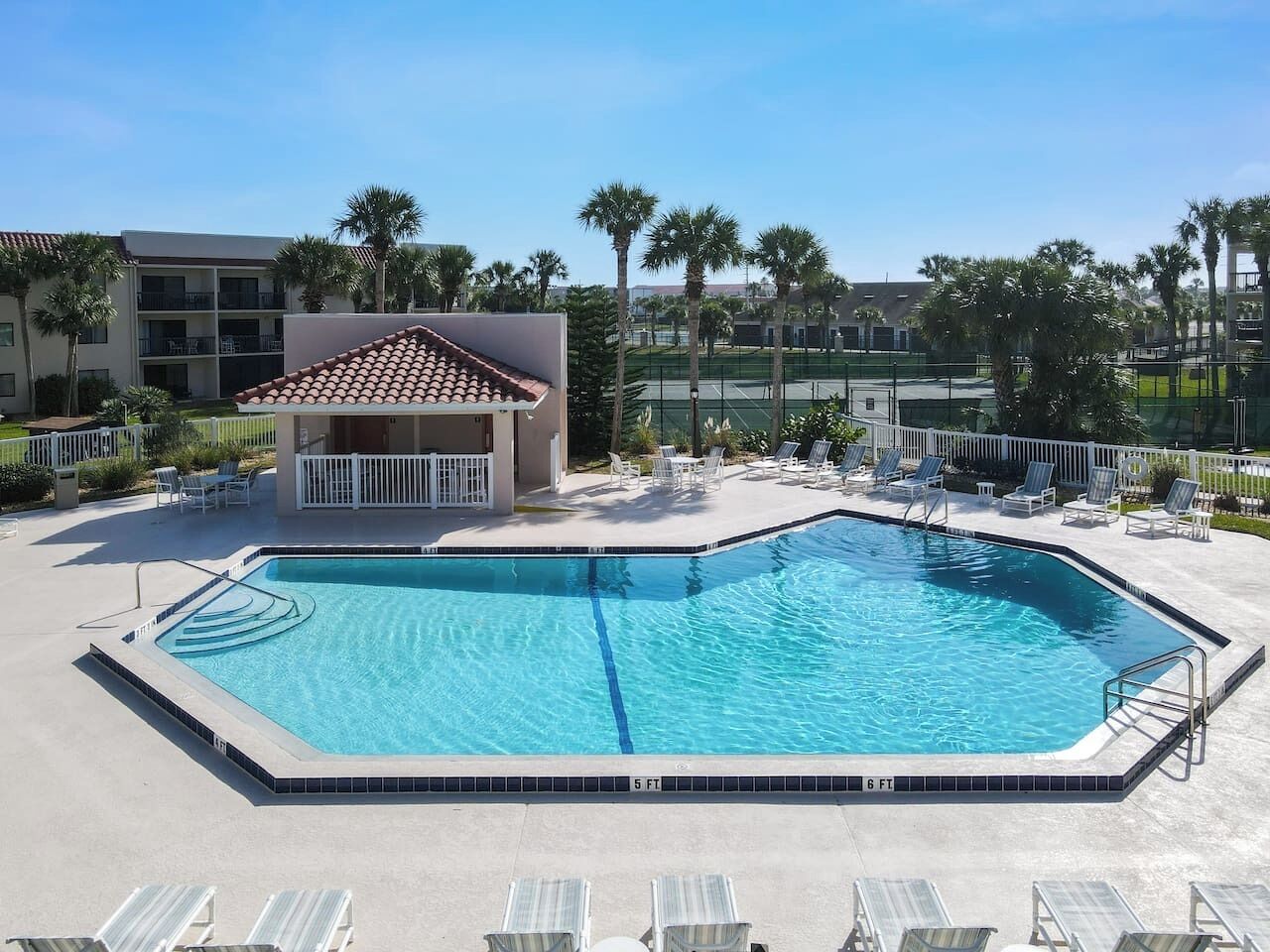 A rectangular swimming pool with a tiled deck, surrounded by lounge chairs and palm trees.