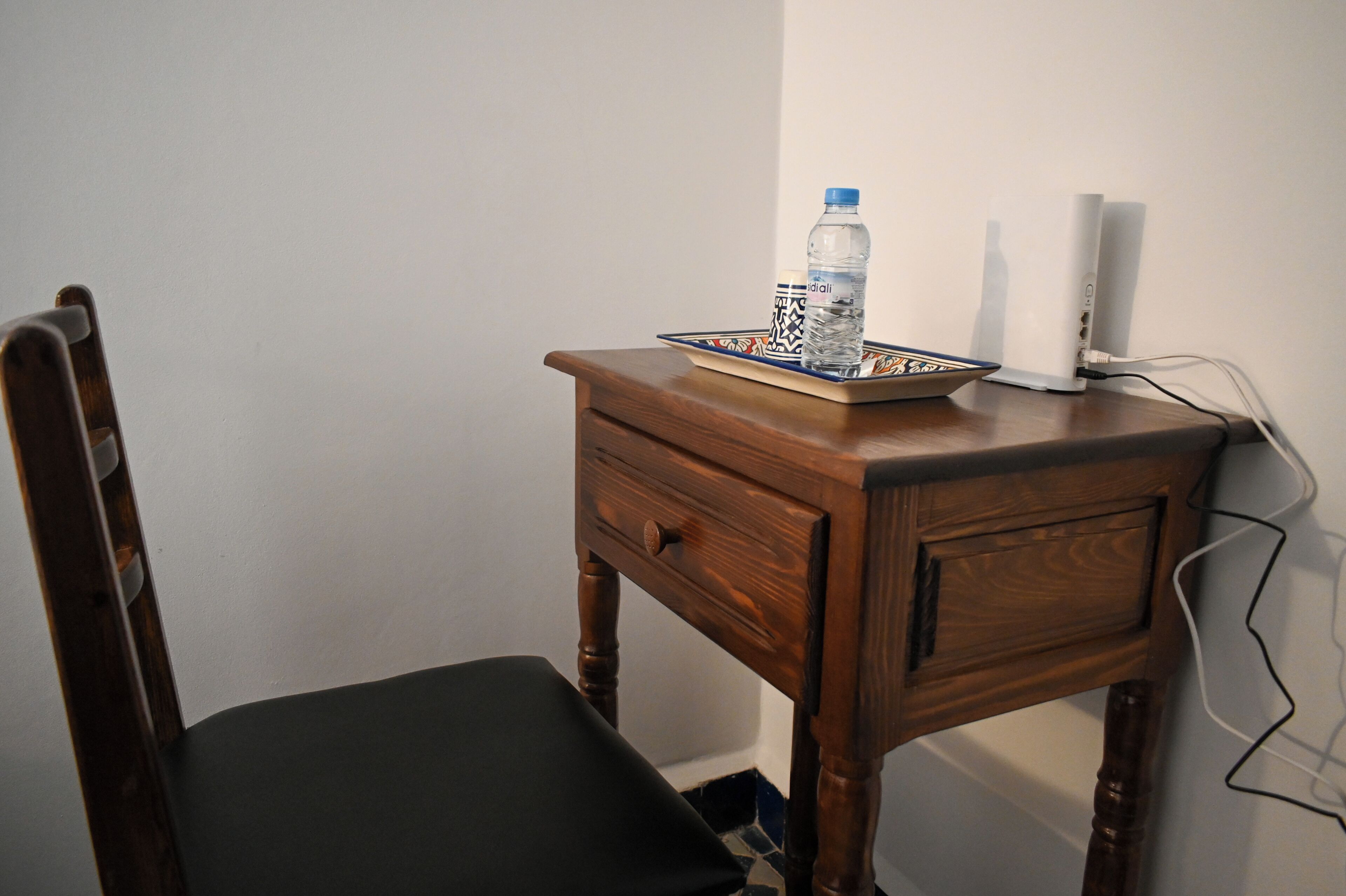 A wooden nightstand with a water bottle, a tray, and a white device, likely a speaker, against a plain wall.