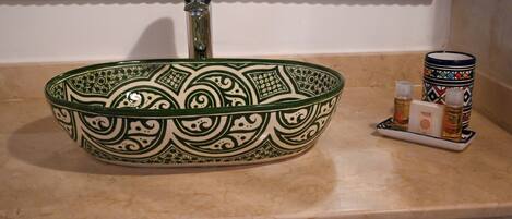 Un lavandino in ceramica verde con decorazioni elaborate, un rubinetto e un piccolo vassoio con due bottiglie su un piano di lavoro color beige.