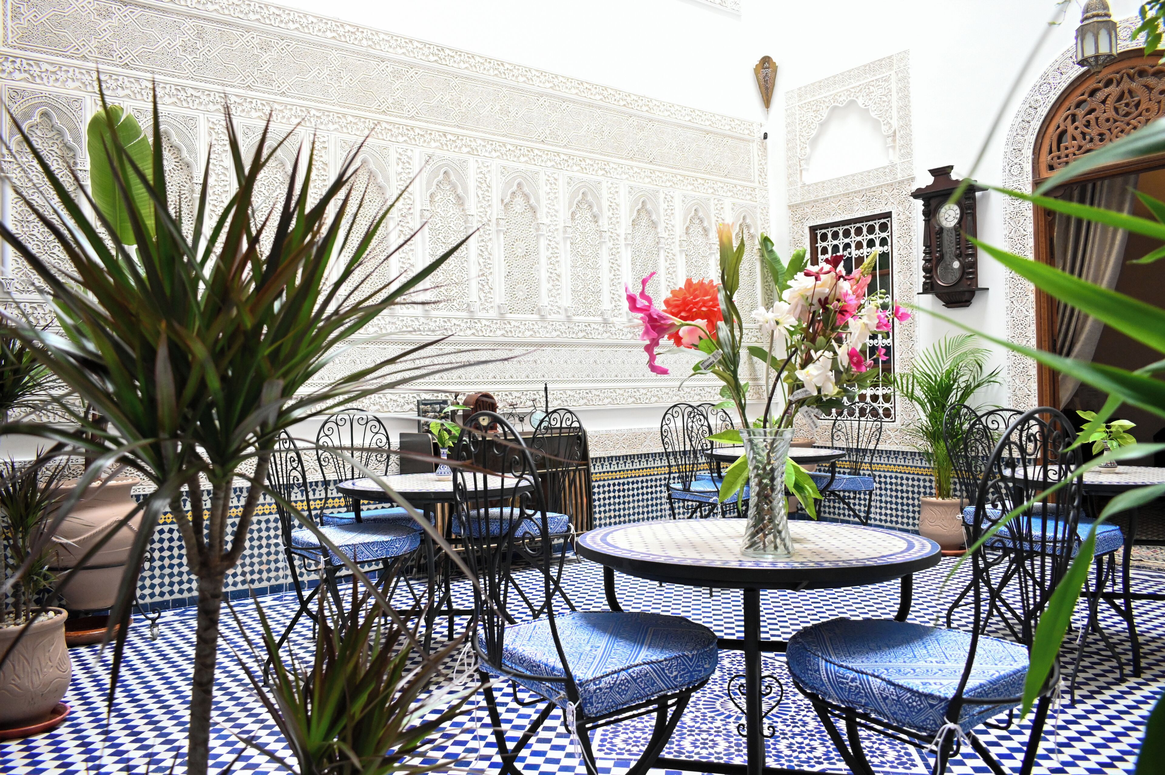 Decoração em estilo marroquino com piso de azulejos estampados, cadeiras com almofadas azuis e paredes brancas ornamentadas.