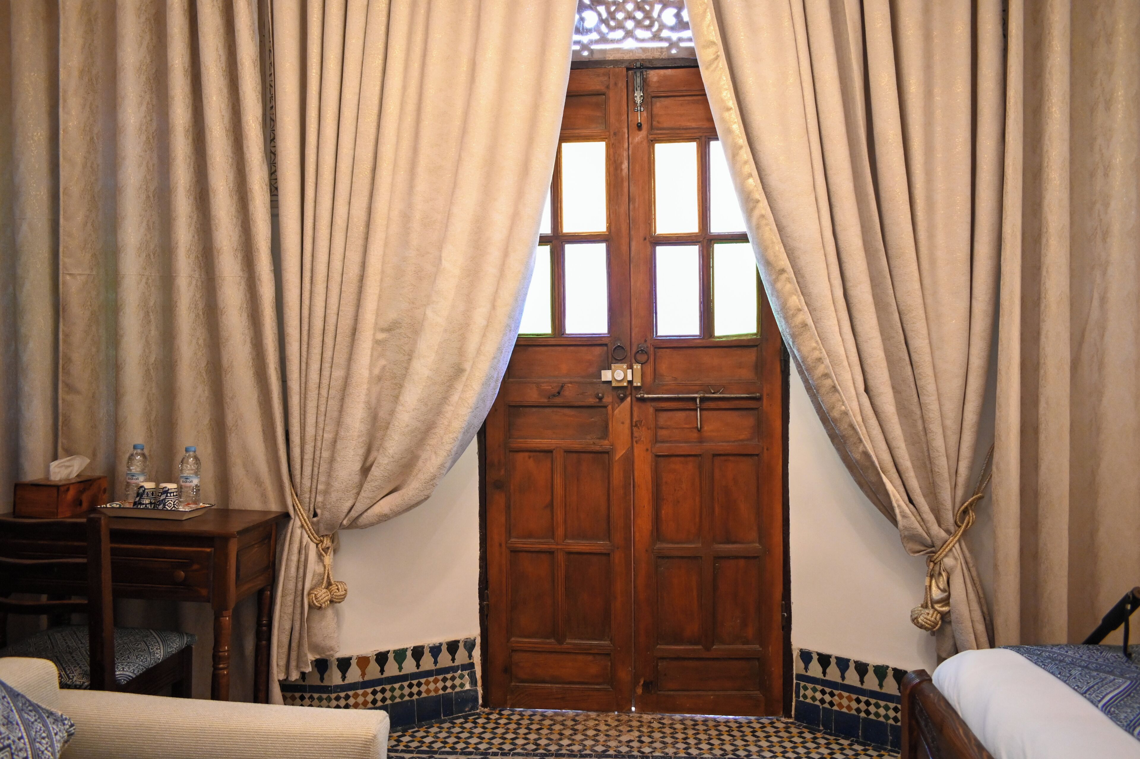 A room with a wooden door, beige curtains, a small table with bottles, and a patterned floor.