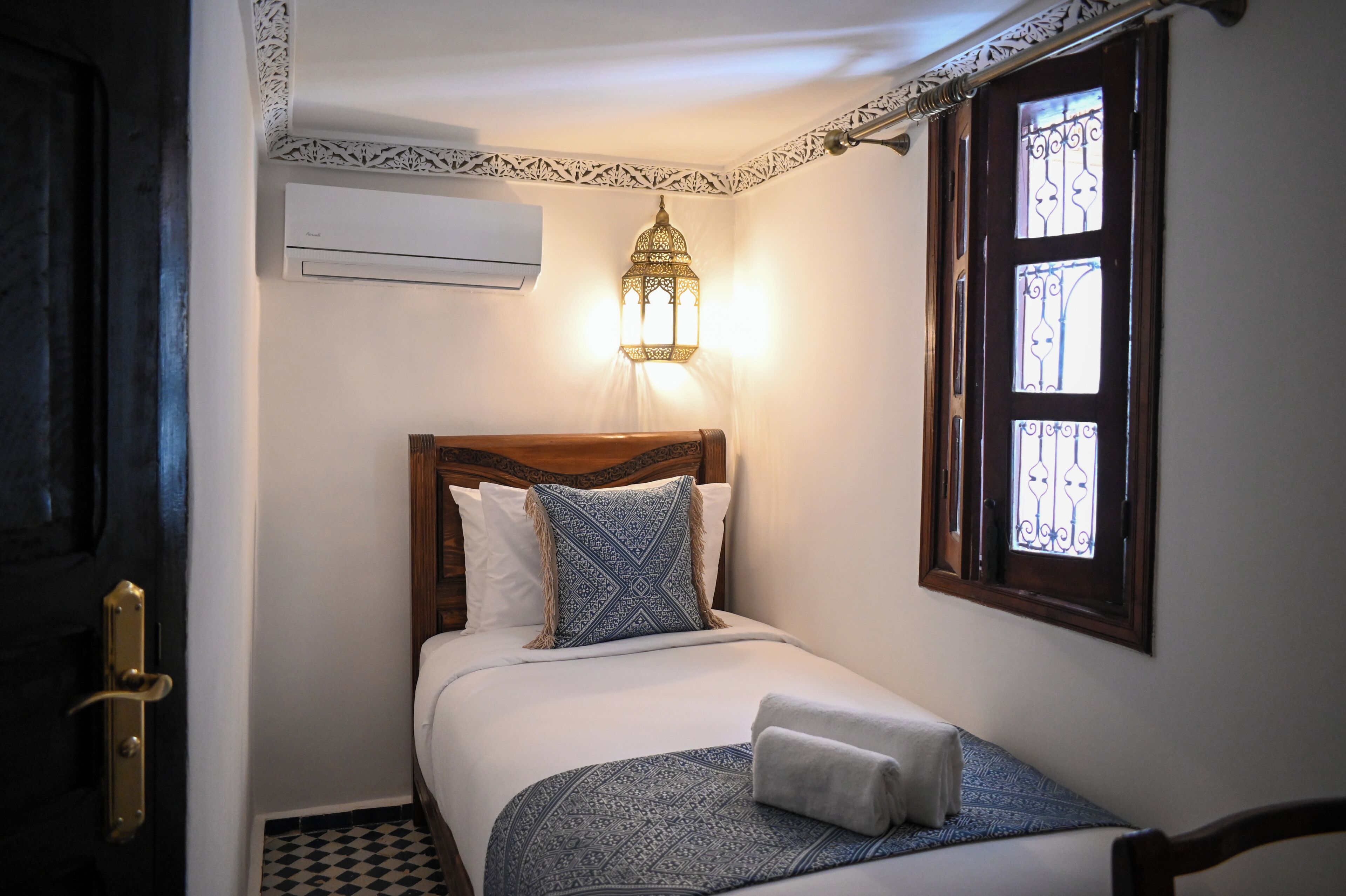 Un lit bien fait, agrémenté d’un coussin à motifs, d’une lampe de chevet et d’une fenêtre ornée de barreaux en fer décoratifs.