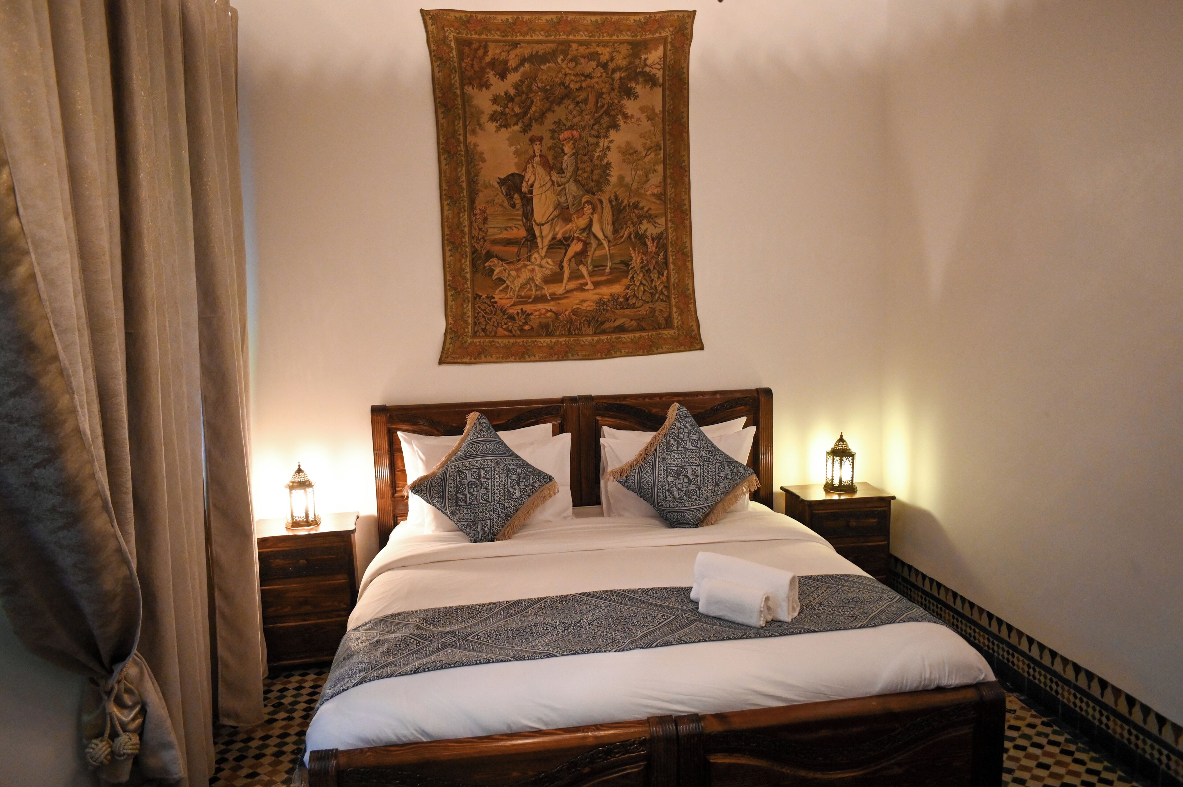A neatly made bed with a patterned bedspread, two decorative pillows, a hanging tapestry, and two bedside lamps.