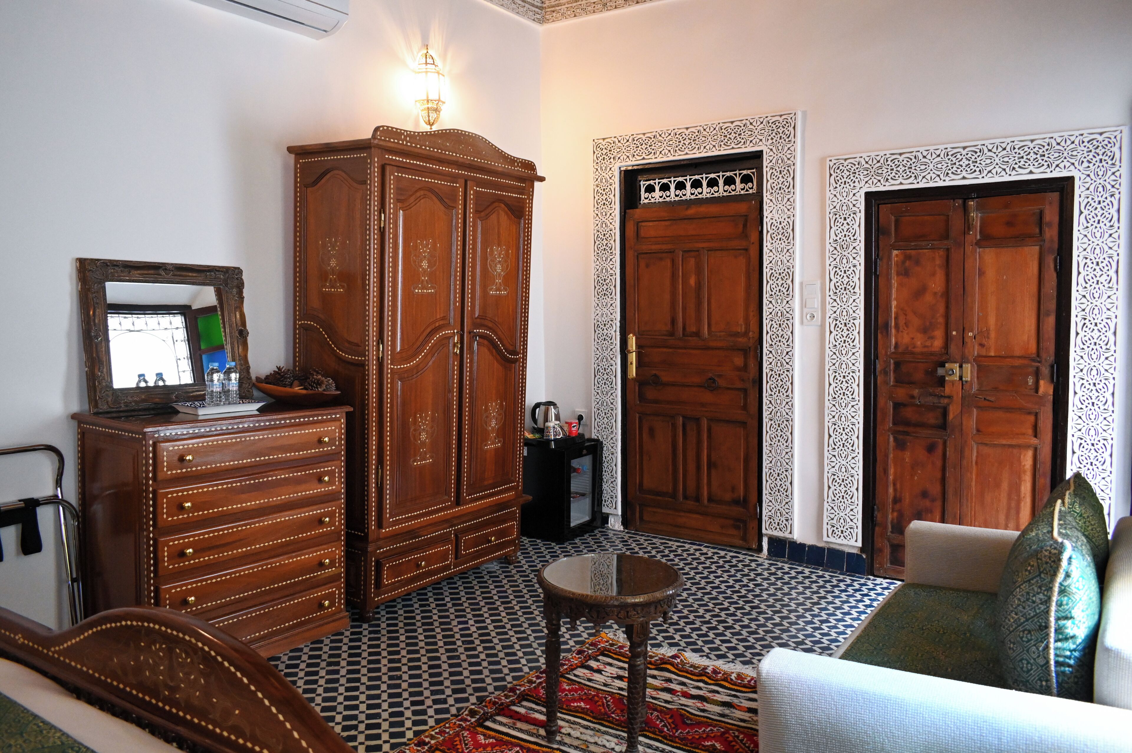 Una habitación con cama, armario de madera, dos puertas de diseño intrincado, una mesita y una alfombra estampada.