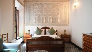 Une chambre avec un mur à motifs, un lit avec des coussins verts, une tête de lit en bois, une table de chevet, une commode et un sol à motifs.