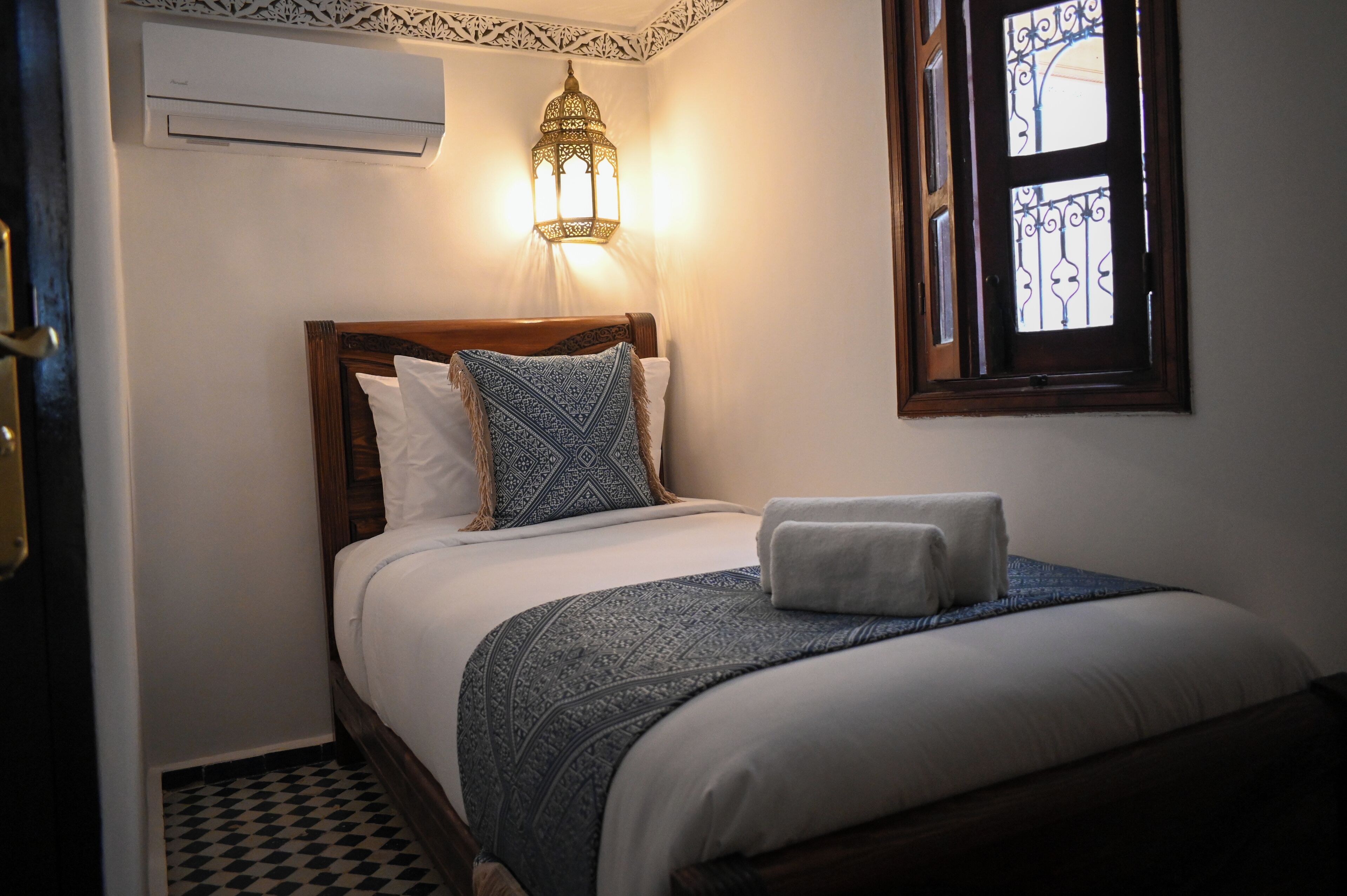 Una cama bien tendida con una colcha estampada, una lámpara de noche y una ventana con barandales de hierro decorativos.