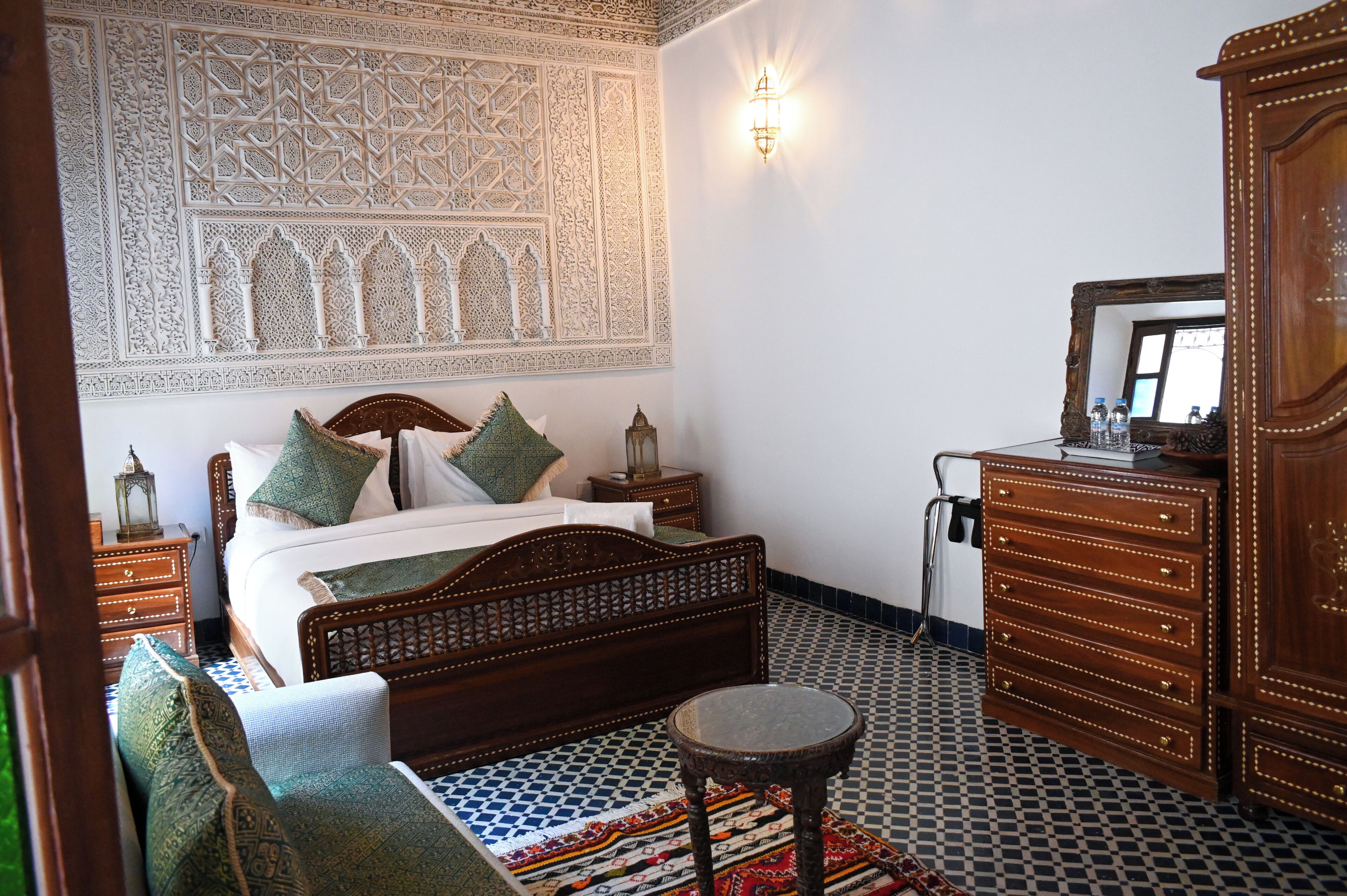 A bedroom with a wooden bed, a nightstand, a dresser, and a wall with intricate patterns.