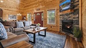 Ein Blockhaushaus-Interieur mit Kamin, einem an einer Steinwand montierten Fernseher und einer gemütlichen Sitzecke mit Sofa und Couchtisch.