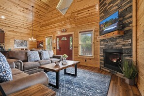 A log cabin interior with a fireplace, a television mounted on a stone wall, and a cozy seating area with a sofa and coffee table. - Gatlinburg*Small but Spacious*WiFi*Hot Tub*Resort Amenities (Gatlinburg)