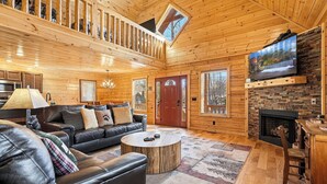 L’intérieur d’une cabane en rondins, avec un canapé, une table basse, une cheminée, un téléviseur et un escalier.