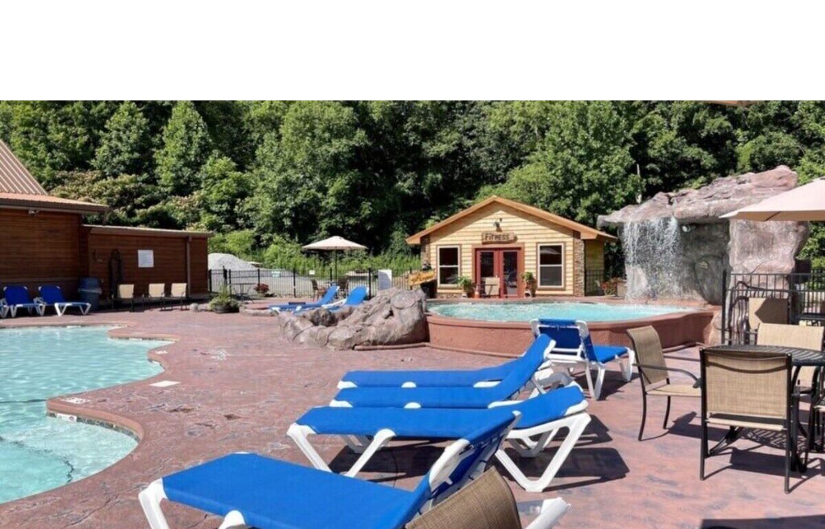 Un espace piscine avec des chaises longues, un petit bâtiment portant l’inscription « Salle de remise en forme » et une cascade décorative.