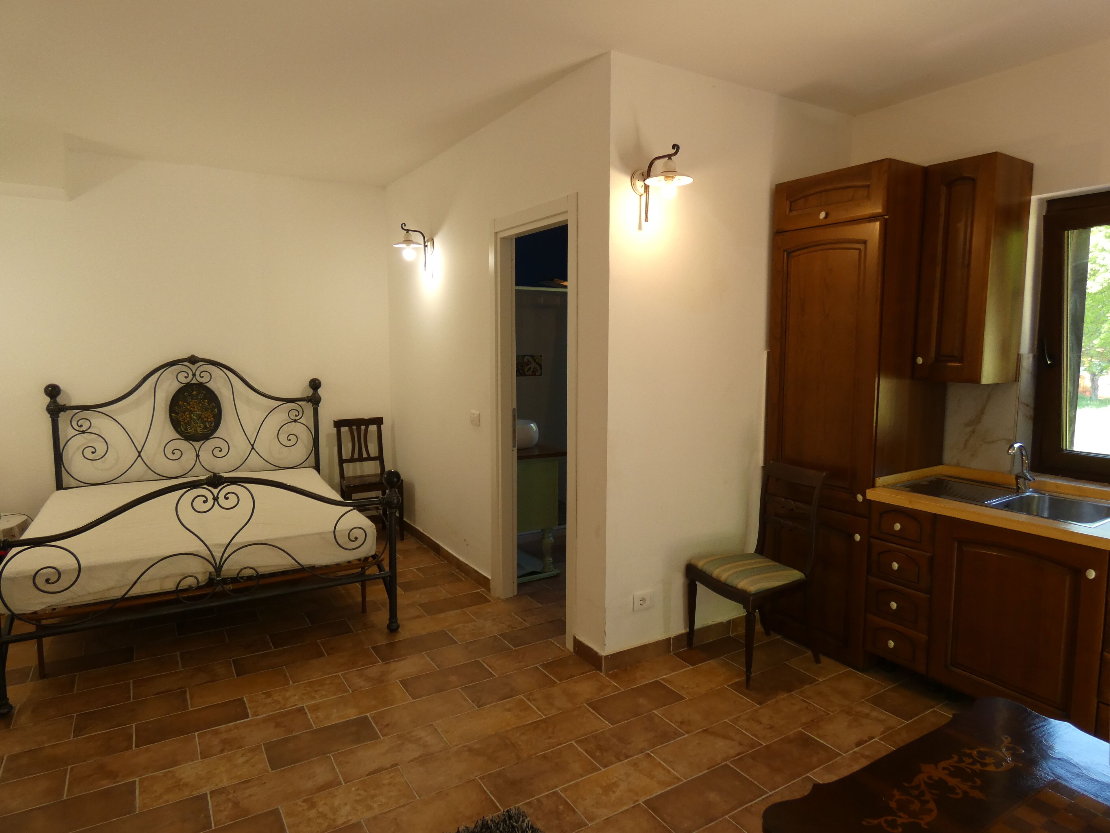 Una camera da letto con un letto, una sedia, un tavolino, una finestra e un angolo cottura con pensili e un lavello.