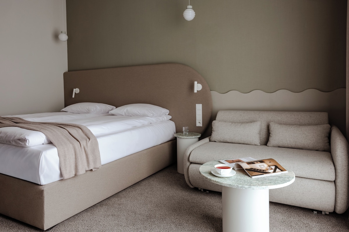 A bedroom with a bed, a sofa, a small table with a book and a cup, and a wall-mounted light fixture.
