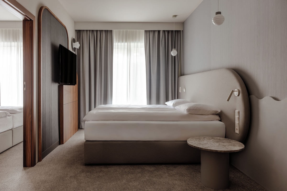 A modern hotel room with a large bed, a small bedside table, a TV mounted on the wall, and a window with curtains.