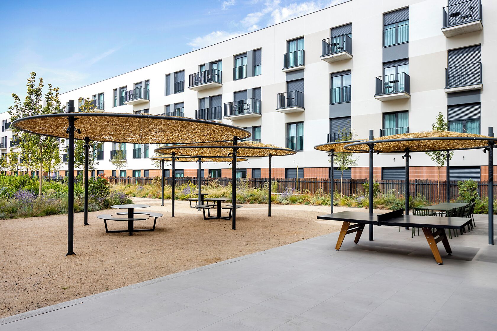 An outdoor patio area with round tables and chairs, covered by woven canopies, in front of a modern apartment building.