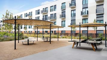 An outdoor patio area with round tables and chairs, covered by woven canopies, in front of a modern apartment building.
