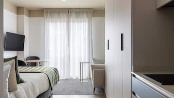 A modern hotel room with a bed, a small round table, a sofa, a TV mounted on the wall, and a large window with sheer curtains.