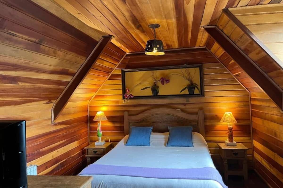 Un dormitorio acogedor en el ático, con paredes de madera, una cama con almohadas azules, dos lámparas de noche y un cuadro enmarcado sobre la cama.