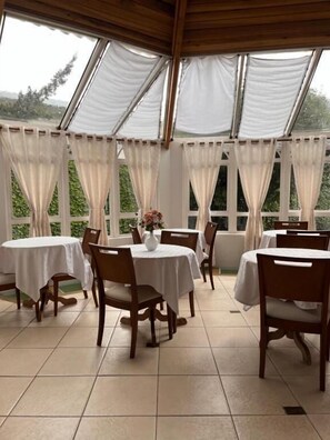 Un área de comedor techada con mesas y sillas, ventanas amplias con cortinas y vistas al exterior con vegetación.