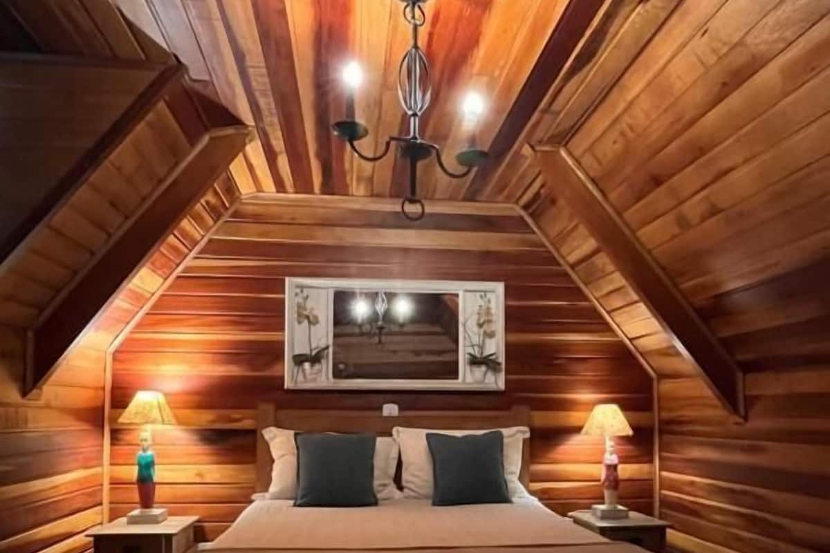 Un dormitorio acogedor en el ático, con paredes de madera, una cama grande, dos lámparas de noche y una araña.