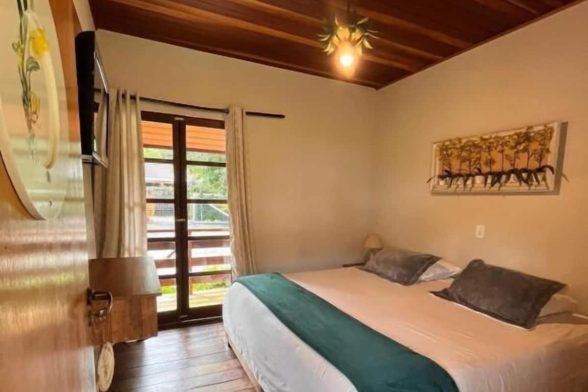 Un dormitorio con techo de madera, cama con ropa de cama blanca y verde, un cuadro en la pared y una ventana con cortinas.