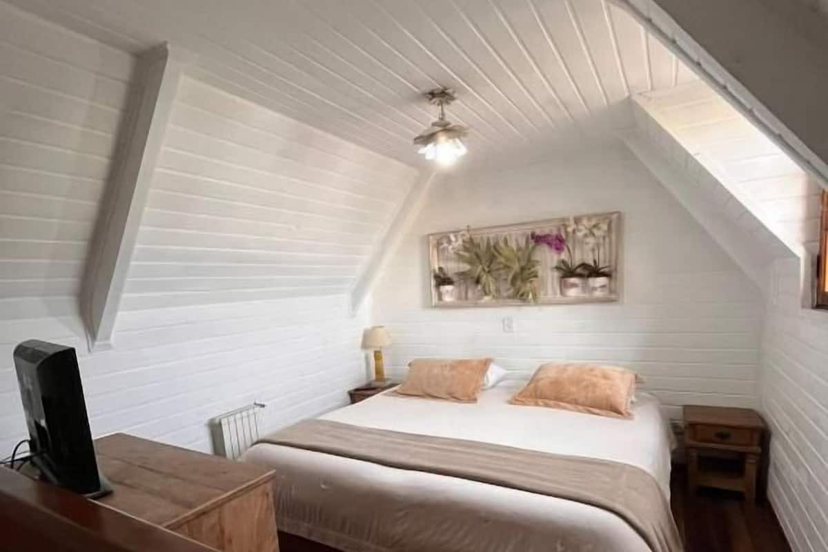 Un dormitorio con techo inclinado, una cama con cabecera, mesitas de noche, un banco de madera y un estante con plantas.