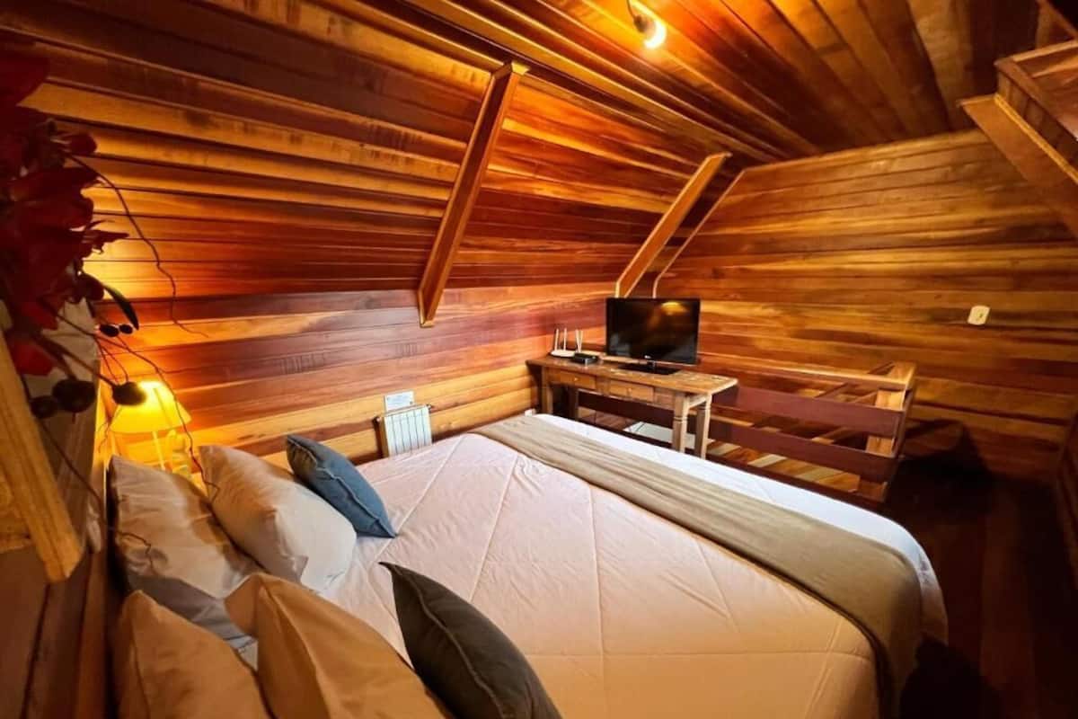 Un dormitorio acogedor en el ático con techo inclinado de madera, una cama con almohadas, una mesita con televisor y una lámpara.