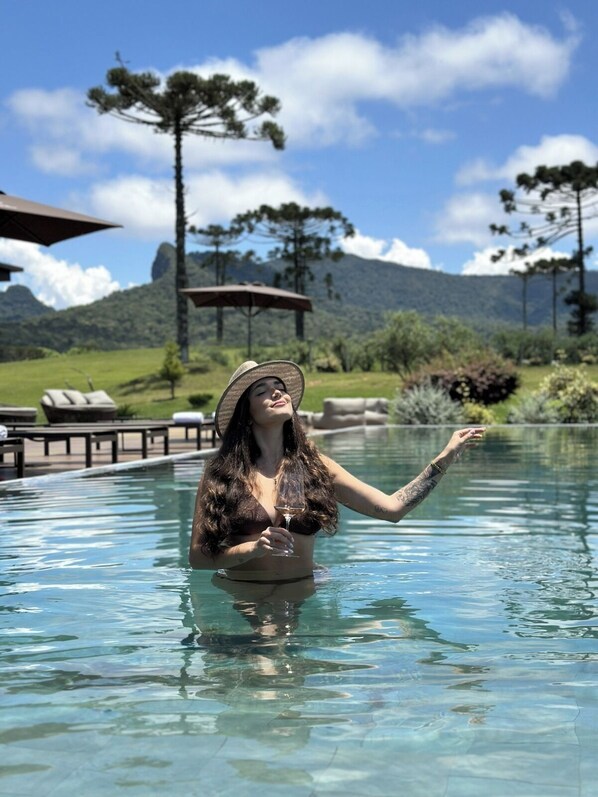A person in a swimming pool with clear water, surrounded by lounge chairs and a mountainous landscape in the background. - Fazenda Bom Retiro Hotel Boutique (Bom Retiro)