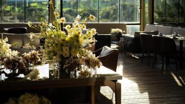 A spacious indoor area with large windows offering a view of mountains and greenery. There is a wooden table with a floral arrangement and a basket of flowers. - Fazenda Bom Retiro Hotel Boutique (Bom Retiro)