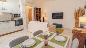 A modern kitchen with white cabinets, a stove, and a microwave. There is a dining area with a table set for two, chairs, and a green placemat. A blue ottoman and a lamp are visible in the background. - Casa Sophie - One Bedroom Apartment, Sleeps 4 (Desenzano del Garda)