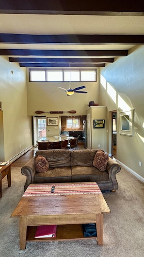 A spacious living room with a high ceiling, a ceiling fan, a dining area, and a kitchen in the background.