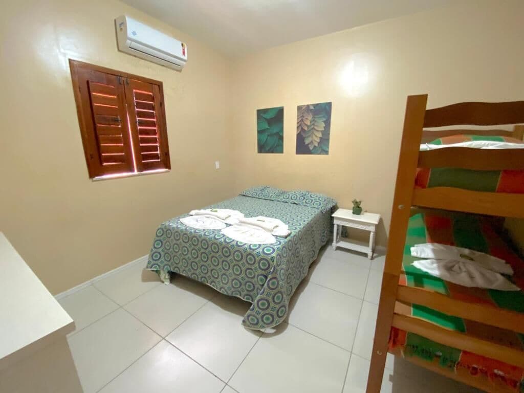 A bedroom with a bed, a small bedside table, a wall-mounted air conditioner, and a window with shutters.