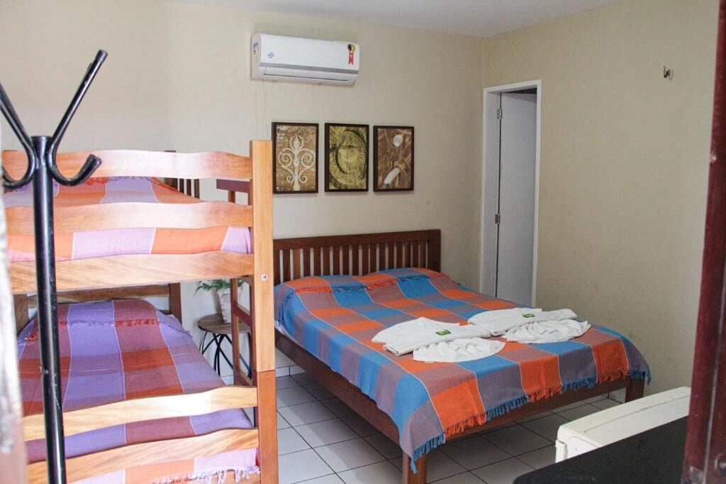 Une chambre avec des lits superposés, un lit recouvert d’une couverture colorée, une petite table avec une plante et trois tableaux encadrés accrochés au mur.