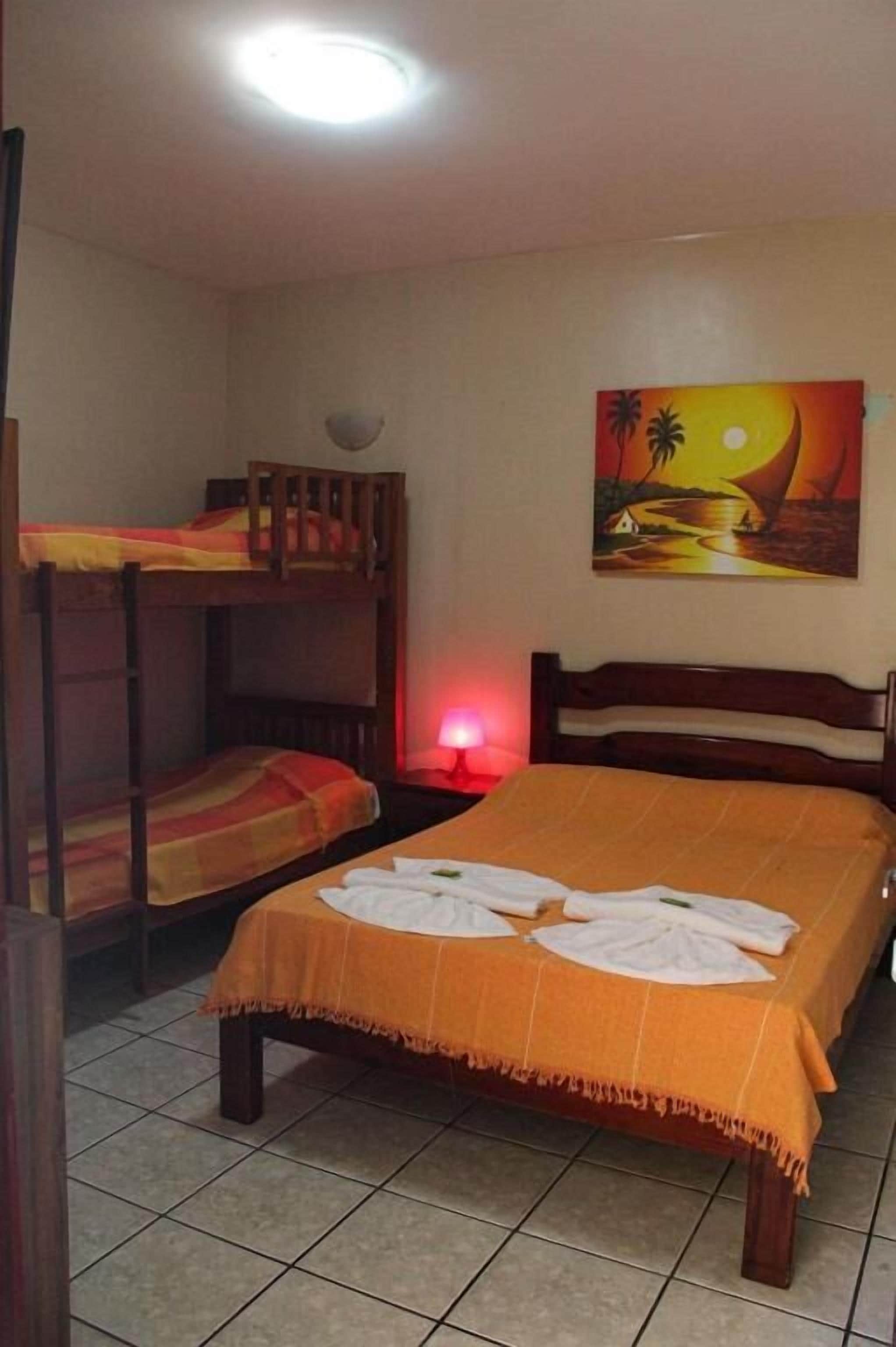 A room with a bunk bed, a single bed with an orange bedspread, a painting of a sunset, and a red lamp on a bedside table.