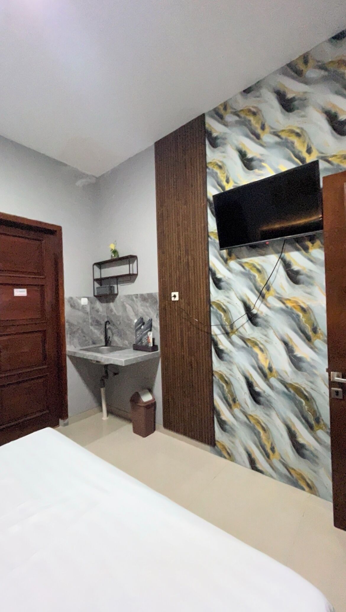 A bedroom with a bed, a wooden wardrobe, a bathroom with a sink and mirror, and a TV mounted on the wall.