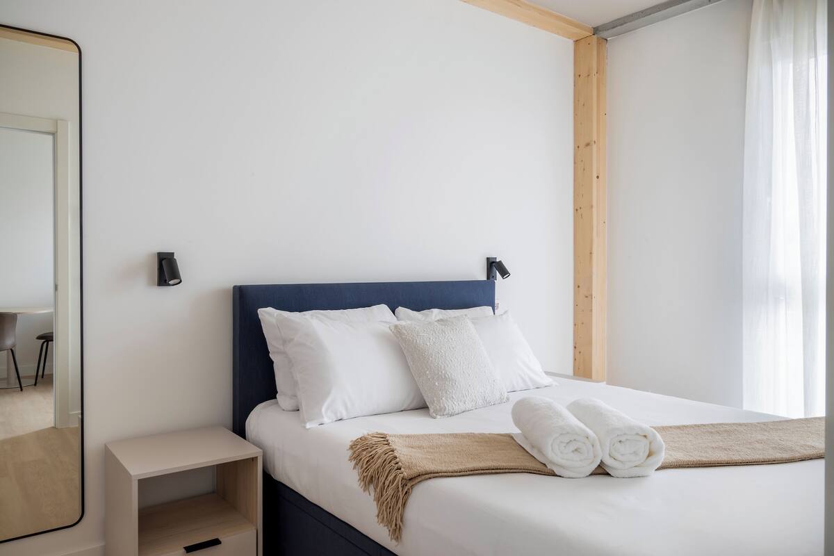 A neatly made bed with white linens and a beige throw blanket, a bedside table with a lamp, and a wooden headboard.