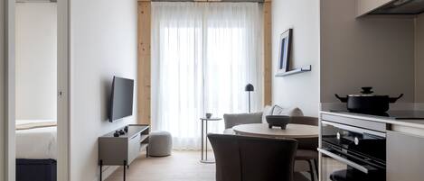 A modern kitchen with a dining area, a TV, and a large window with sheer curtains.