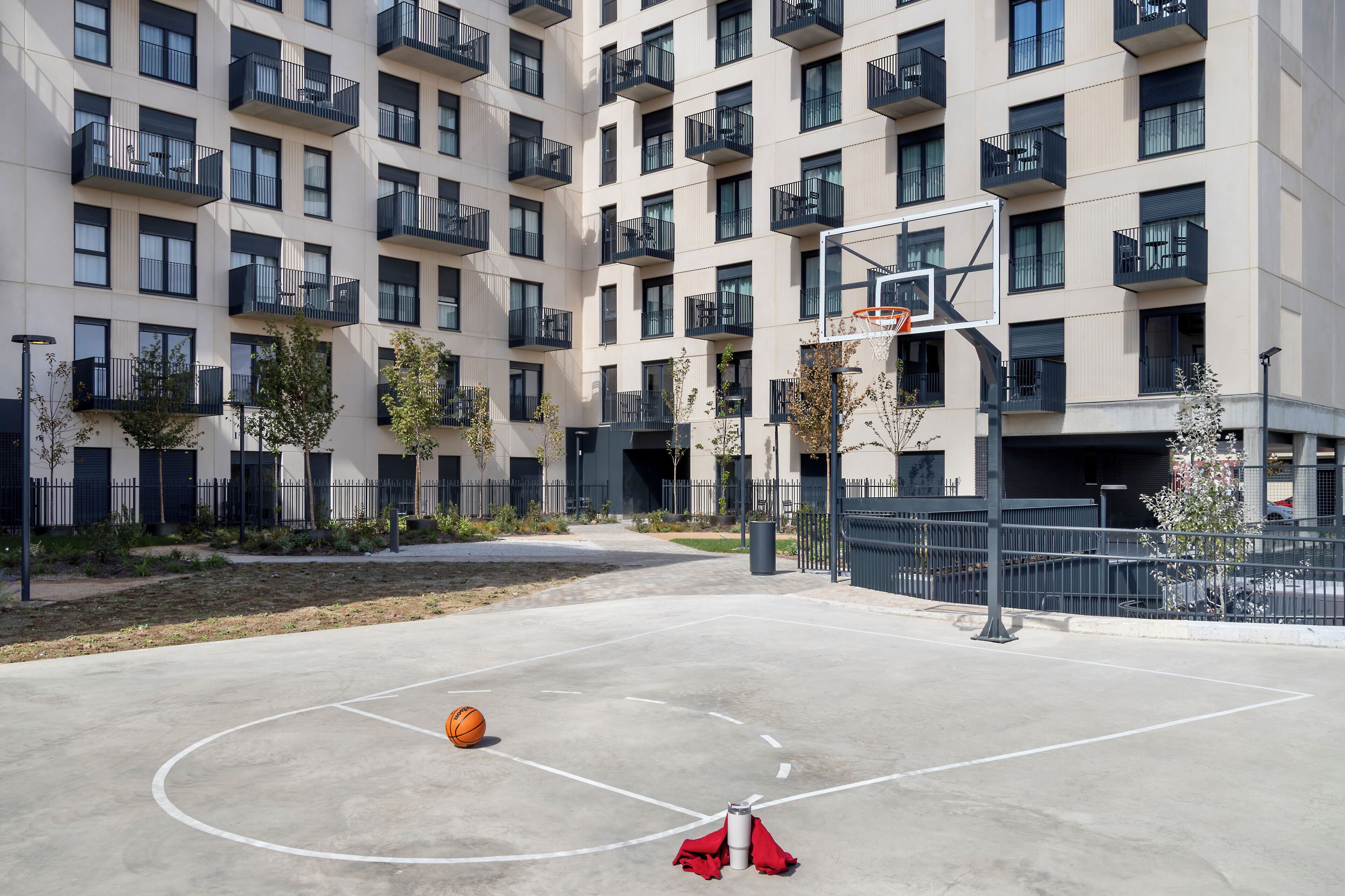 Een buitenbasketbalveld met een basket, omringd door een meerlaags woongebouw met balkons.