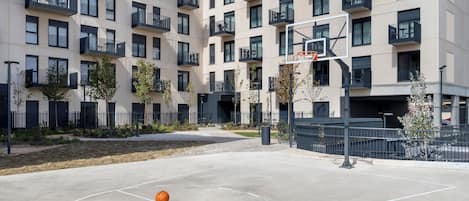 Una cancha de baloncesto al aire libre con un aro, rodeada por un edificio residencial de varios pisos con balcones.