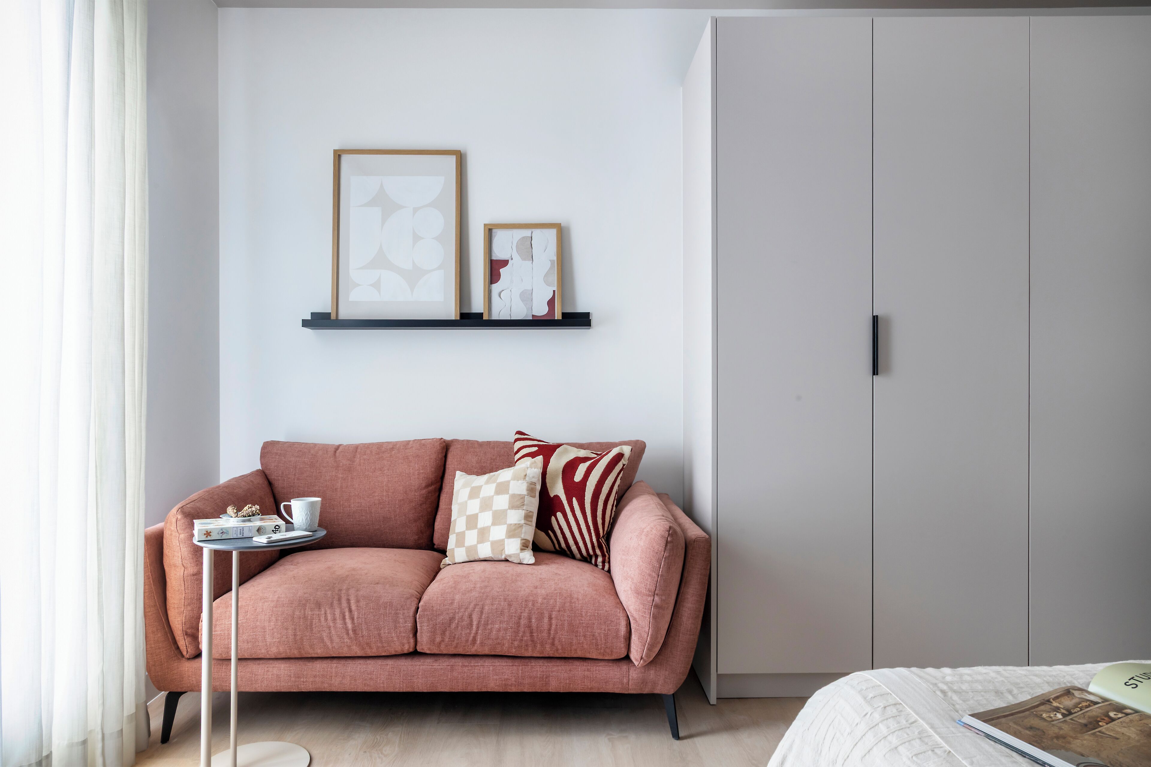 Ett modernt rum med en rosa soffa, ett litet sidobord med ett bricka på och en säng med ett vitt överkast. På väggen ovanför soffan hänger två inramade, abstrakta konstverk.