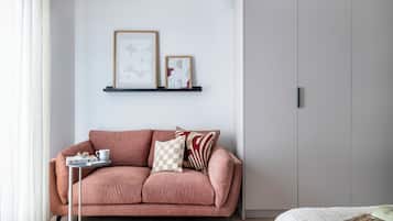 Ett modernt rum med en rosa soffa, ett litet sidobord med ett bricka på och en säng med ett vitt överkast. På väggen ovanför soffan hänger två inramade, abstrakta konstverk.