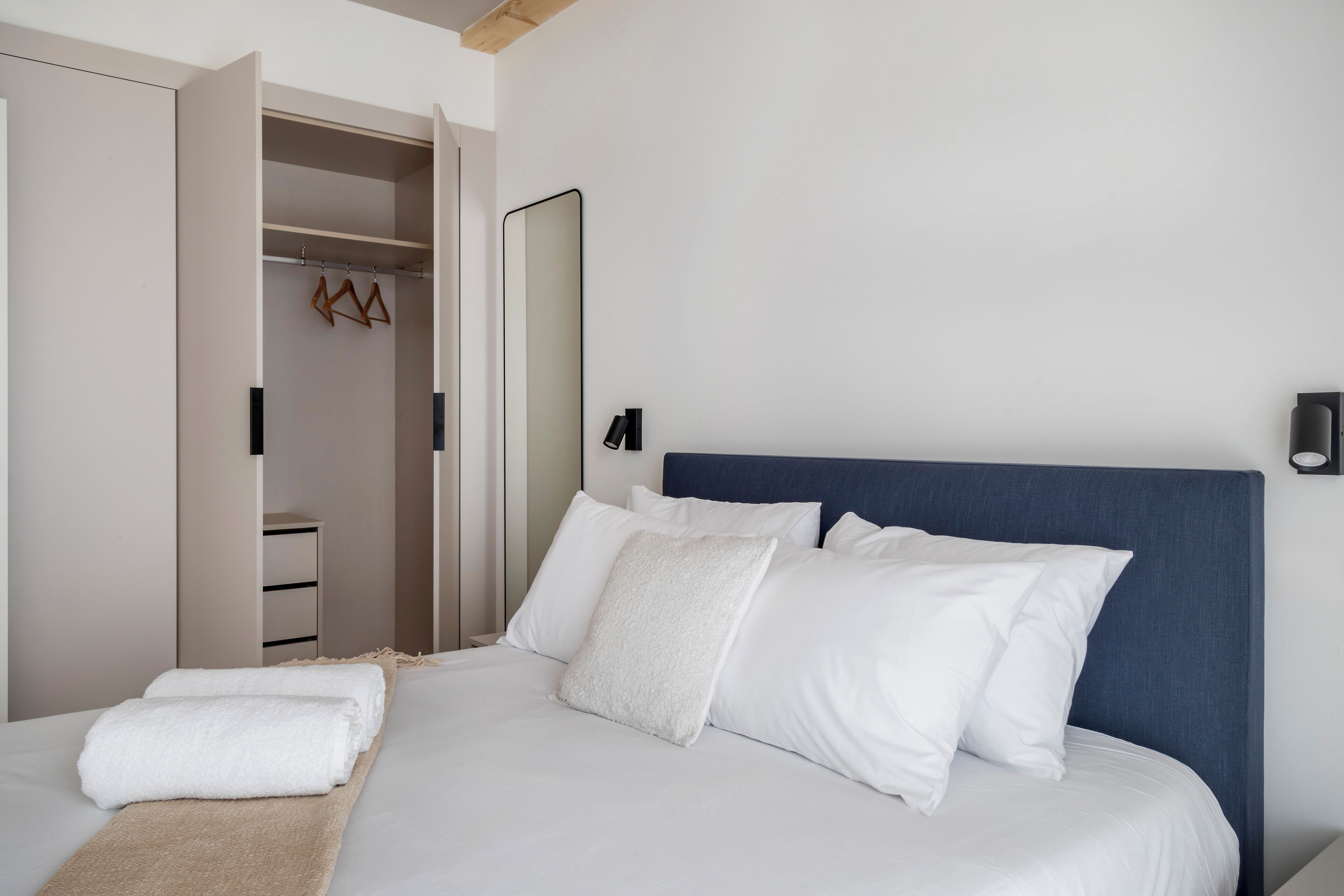 A neatly made bed with white linens and a blue headboard, a wooden hanger rack, and a mirror on the wall.
