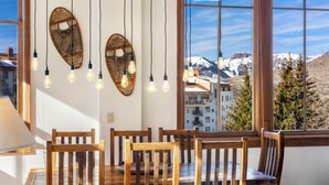 Et spiseområde med træstole og et bord, en hængende lampe og udsigt til sneklædte bjerge gennem store vinduer.