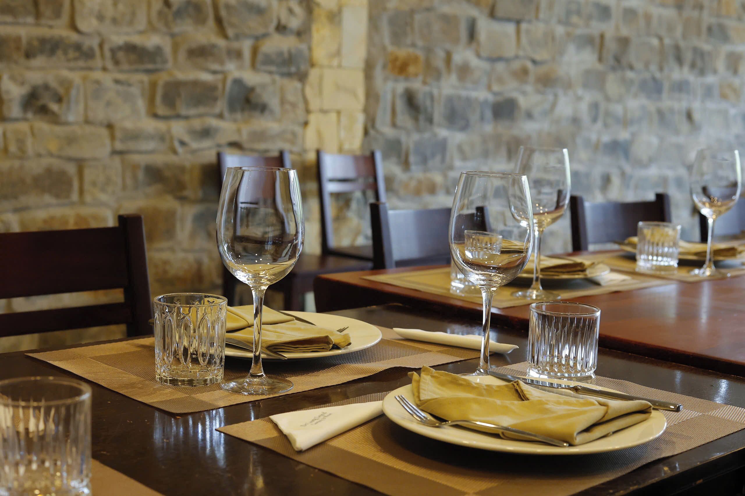 A dining table set with wine glasses, plates, and cutlery, with a stone wall in the background.