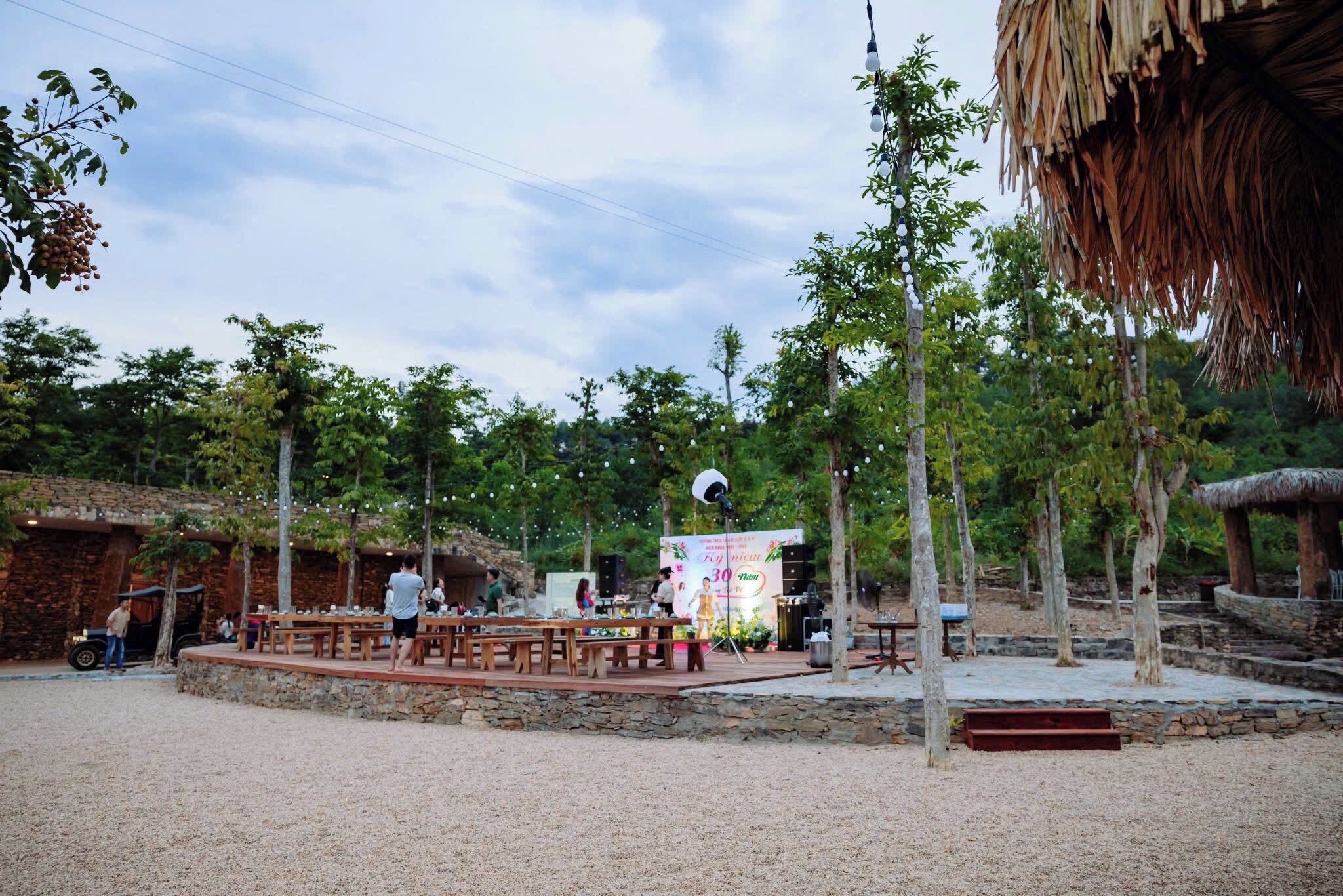 An outdoor dining area with wooden tables and benches, surrounded by trees and a thatched-roof structure.